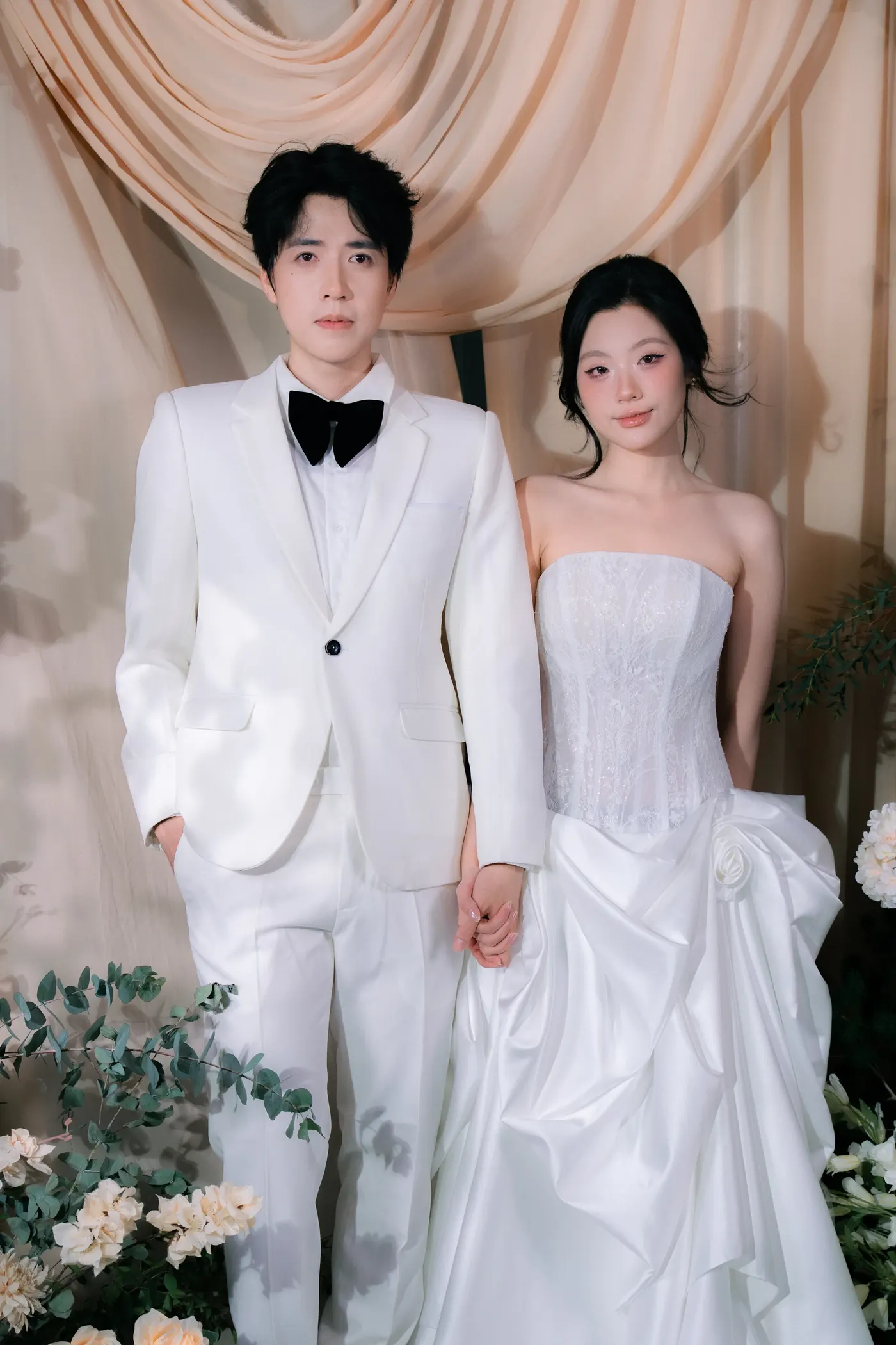 Aiden and Paige in white wedding attire in front of draped ivory fabric