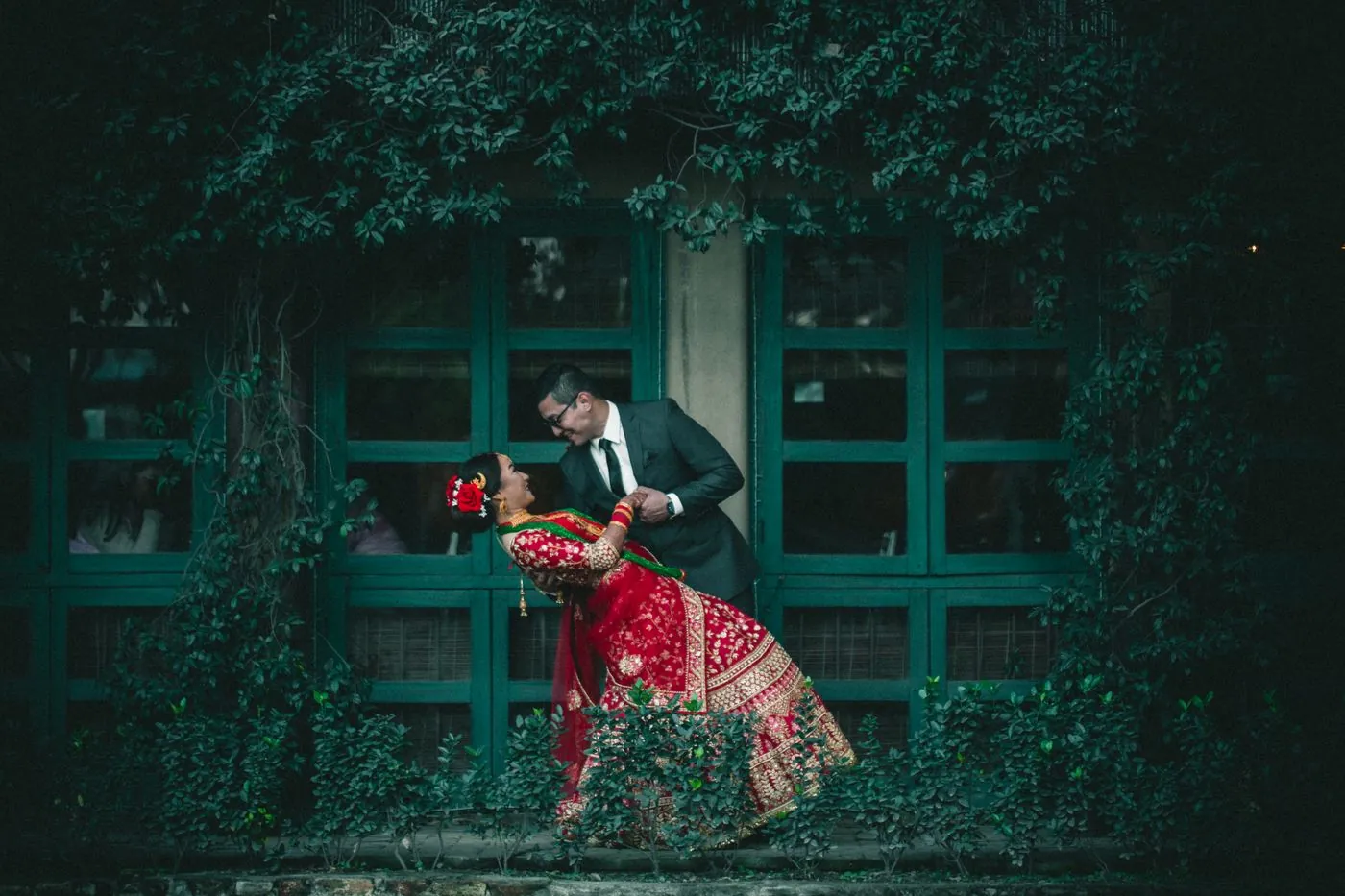 A bride in a red lehenga and a groom in a suit sharing a dip in front of an ivy-covered wall