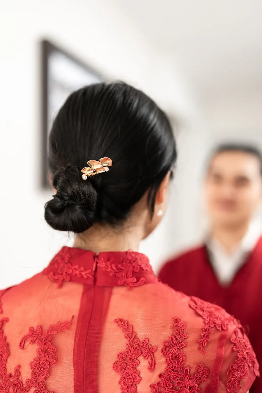 The back of a bride in a red lace qipao with a gold hairpin, looking toward the groom
