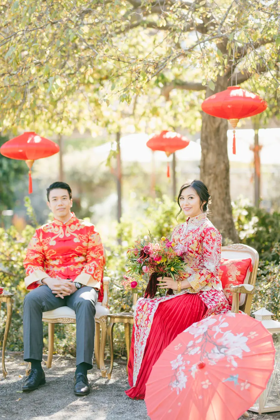 A couple in traditional red Chinese wedding attire seated with a red parasol under lanterns